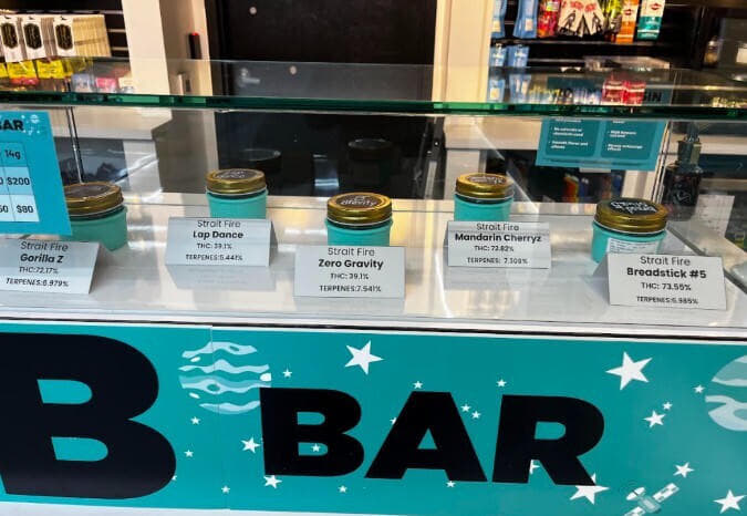 A glass display case at a cannabis dispensary showcasing five jars of flower labeled “Strait Fire.” The strains visible are Gorilla Z, Lap Dance, Zero Gravity, Mandarin Cherryz, and Breadstick #5, each with their THC and terpene percentages listed on small cards. The counter features a teal design with stars and the word “BAR” in bold black letters.